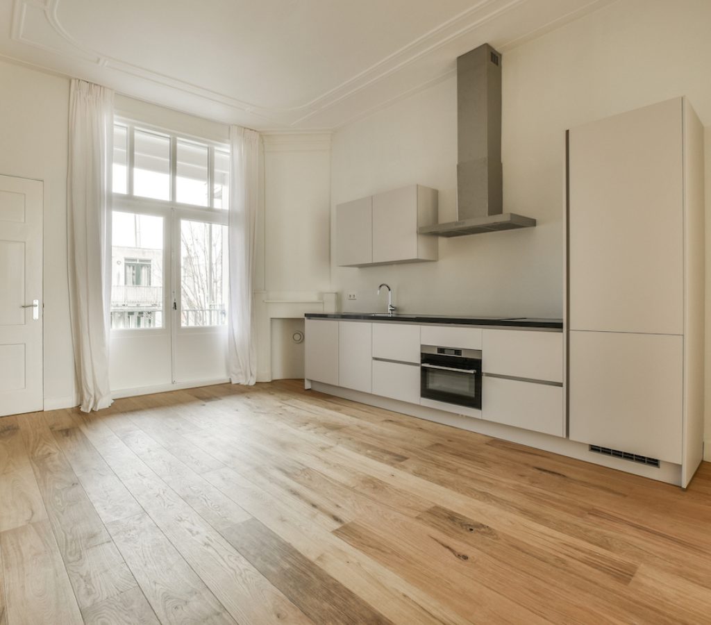 an empty kitchen with wood flooring and white appliances in the middle of the room, there is a window that looks out to
