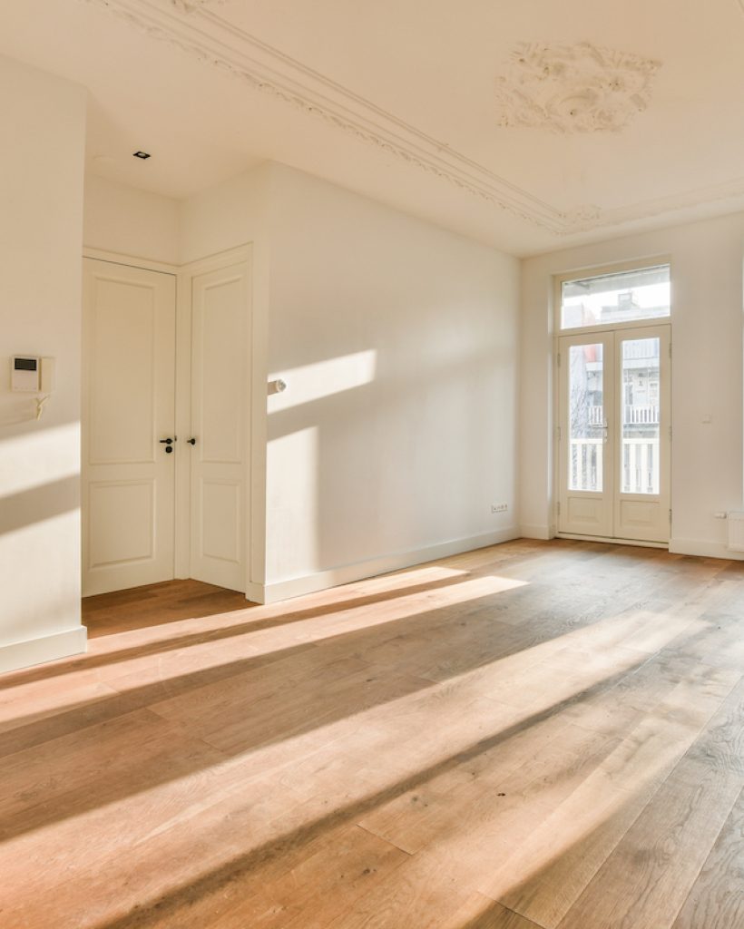 an empty living room with wood flooring and white painted ceiling in the background is sunlight shining through two large windows